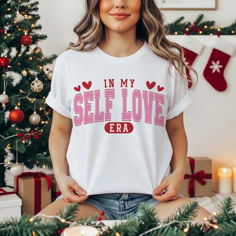 May include: White t-shirt with the text "IN MY SELF LOVE ERA" in pink and red. The shirt has a crew neck and short sleeves. The model is wearing denim shorts. The background includes a Christmas tree and decorations.