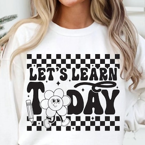 Op de afbeelding: Witte sweatshirt met een zwart-wit geblokt patroon aan de boven- en onderkant. De afbeelding in het midden zegt "LET'S LEARN TODAY" met een cartoon bloem en potlood illustratie.