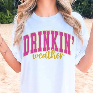May include: White t-shirt with the text "DRINKIN' weather" in pink and gold letters.