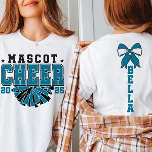 May include: White t-shirt with the text "MASCOT CHEER 20 25" and a blue and black cheerleading pom-pom graphic. The back of the shirt has a blue bow with the text "BELLA" written vertically.