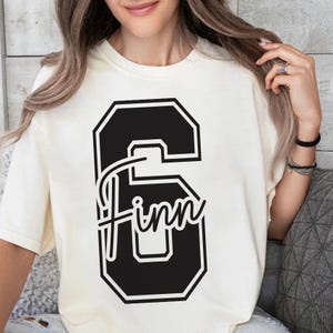 May include: Cream-colored t-shirt featuring a large black number 6 with the name "Finn" in a script font. The design is centered on the front of the shirt. The shirt has a crew neck and short sleeves.