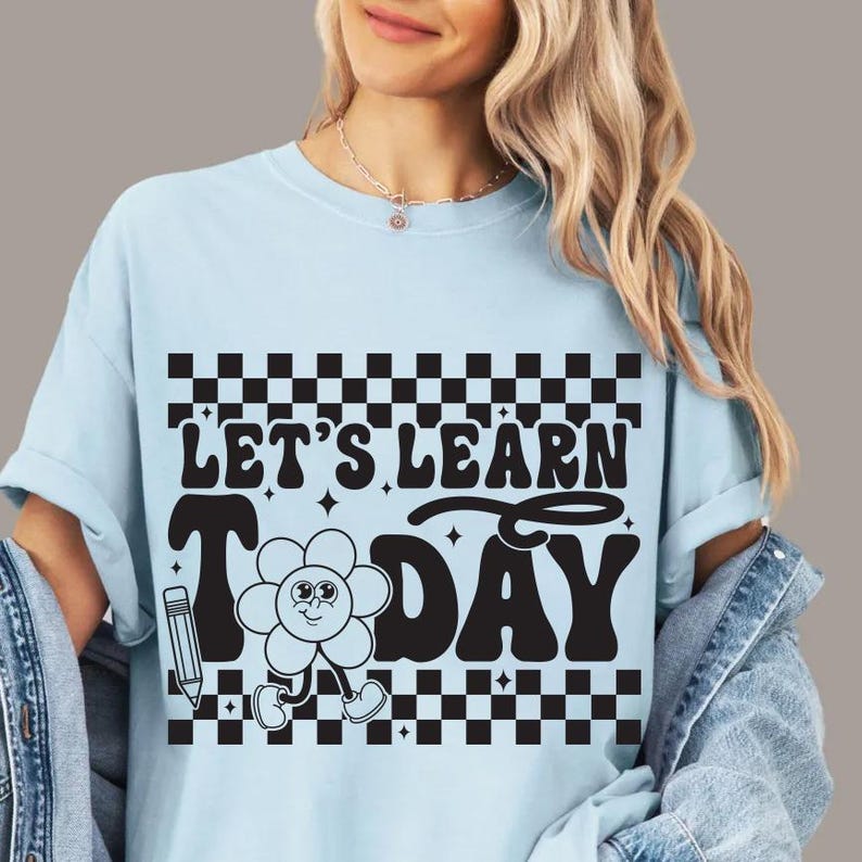 Op de afbeelding: Lichtblauw T-shirt met de zwarte tekst "LET'S LEARN TODAY", met een bloemfiguur en een potlood. Het ontwerp bevat een zwart-wit geblokt patroon. Het shirt is een casual kledingstuk.