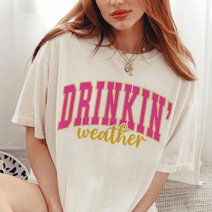 May include: White t-shirt with a pink and gold graphic that says "Drinkin' weather".