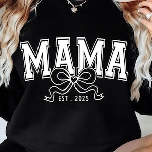Op de afbeelding: Zwart sweatshirt met witte tekst "MAMA EST. 2025" met een witte strik en een hartgrafiek.