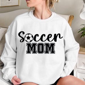 May include: White sweatshirt with black text that reads "Soccer Mom" and a black and white soccer ball graphic.