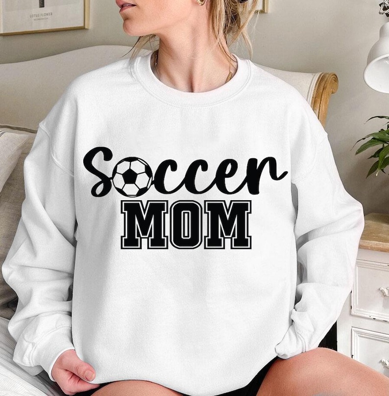 May include: White sweatshirt with black text that reads "Soccer Mom" and a black and white soccer ball graphic.