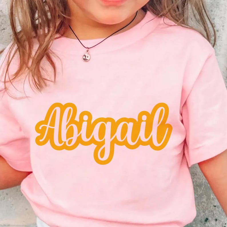 K&ouml;nnte beinhalten: Hellrosa T-Shirt mit dem Namen "Abigail" in einem Retro-Orange-Schriftzug. Das T-Shirt hat kurze &Auml;rmel. Eine kleine Anh&auml;nger-Halskette ist am Ausschnitt zu sehen. Das T-Shirt wird von einer Person getragen.