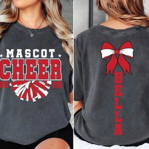May include: A gray t-shirt with the text "Mascot Cheer 2023" and a red and white pom-pom graphic on the front. The back of the shirt has a red bow with white accents and the name "Bella" written vertically in red.