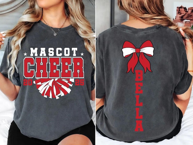 May include: A gray t-shirt with the text "Mascot Cheer 2023" and a red and white pom-pom graphic on the front. The back of the shirt has a red bow with white accents and the name "Bella" written vertically in red.