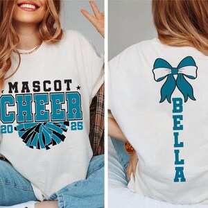 May include: White t-shirt with a blue and black cheerleading pom-pom graphic and the text "Mascot Cheer 20 25". The back of the shirt has a blue bow and the text "Bella" vertically down the back.