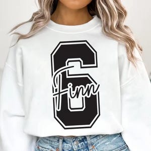May include: White sweatshirt featuring a large black number 6 with a white outline. The name "Finn" is written in a cursive font across the number. The sweatshirt is a casual style.