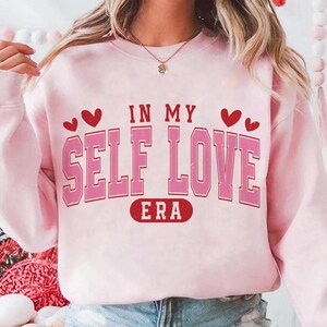 May include: A light pink sweatshirt with the text "IN MY SELF LOVE ERA" in a pink, outlined font. Two red hearts are on each side of the text. The sweatshirt is a casual style.