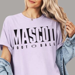 May include: A light purple t-shirt with the word "MASCOT" in large black block letters, with "FOOTBALL" below. A football graphic is between the two words. The shirt is a short-sleeved crew neck.