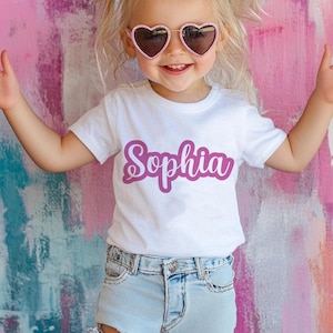 K&ouml;nnte beinhalten: Ein wei&szlig;es T-Shirt mit dem Namen "Sophia" in einer magentafarbenen Schreibschrift. Das Kind tr&auml;gt herzf&ouml;rmige Sonnenbrillen mit rosa Rahmen. Der Hintergrund ist eine farbenfrohe Mischung aus Rosa, Lila und Wei&szlig;.