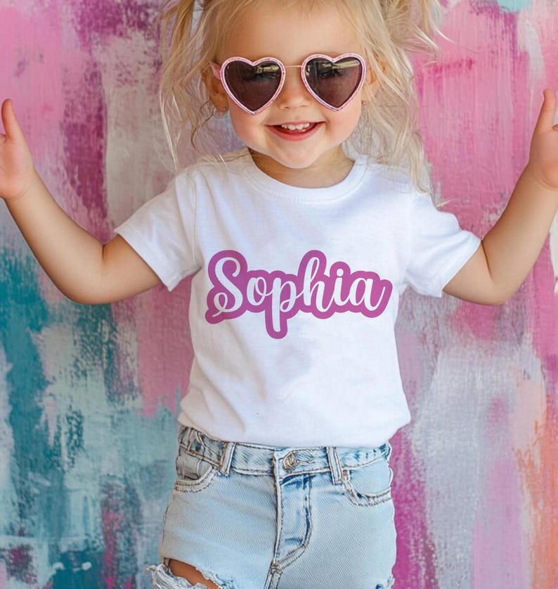 K&ouml;nnte beinhalten: Ein wei&szlig;es T-Shirt mit dem Namen "Sophia" in einer magentafarbenen Schreibschrift. Das Kind tr&auml;gt herzf&ouml;rmige Sonnenbrillen mit rosa Rahmen. Der Hintergrund ist eine farbenfrohe Mischung aus Rosa, Lila und Wei&szlig;.