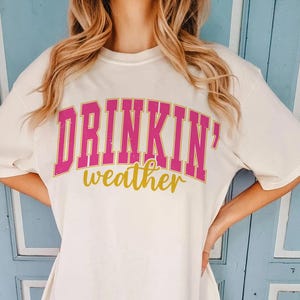 May include: White t-shirt with pink and gold lettering that reads "Drinkin' weather".