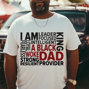 Op de afbeelding: Wit T-shirt met de tekst "I AM A BLACK DAD" in zwart en rood. Extra woorden zijn "Leader, Focused, Intelligent, Fearless, Loyal, Woke, Strong, Resilient, Provider, King". Ronde hals.