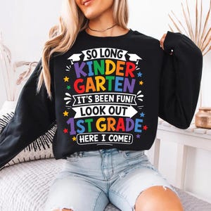 May include: Black long-sleeve sweatshirt with the text "SO LONG KINDERGARTEN IT'S BEEN FUN! LOOK OUT 1ST GRADE HERE I COME!" in colorful lettering. The design includes stars and a banner. A fun, celebratory garment for a child.