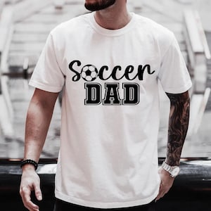 May include: White t-shirt with black text that reads "Soccer Dad" and a black and white soccer ball graphic.