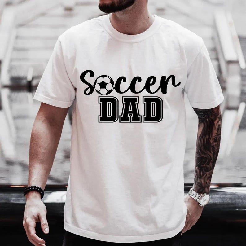 May include: White t-shirt with black text that reads "Soccer Dad" and a black and white soccer ball graphic.