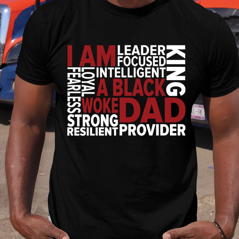 Op de afbeelding: Zwart T-shirt met de rode woorden "I AM", gevolgd door een lijst met positieve eigenschappen in het wit, en de rode woorden "A BLACK DAD" en de witte woorden "KING". Ronde hals.