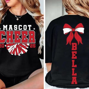 May include: Black cheerleading shirt with red and white pom-poms and the text "Mascot Cheer 2025". The back of the shirt has a red bow and the name "Bella" written vertically.