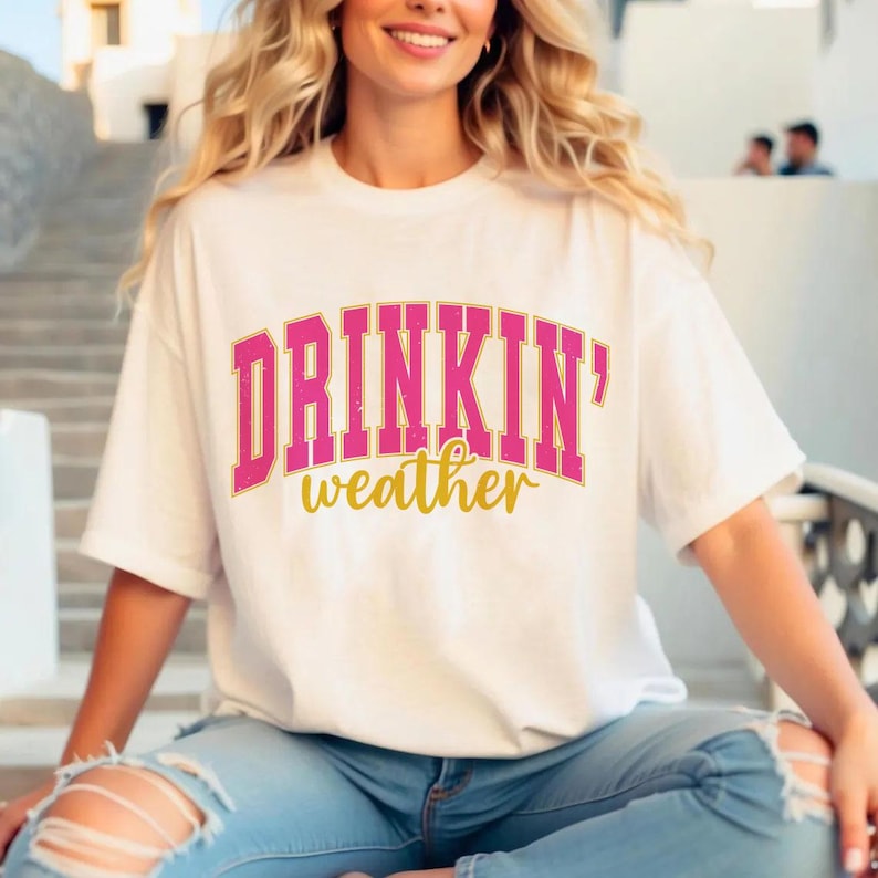 May include: White t-shirt with a pink and gold graphic that says "DRINKIN' weather".