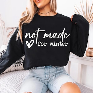 May include: Black sweatshirt with the white text "not made for winter" and a heart symbol. The long-sleeved top is a solid black color. The sweatshirt is a casual style.