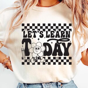 Op de afbeelding: Cr&egrave;mekleurig T-shirt met de tekst "LET'S LEARN TODAY" in een retro lettertype. Het ontwerp bevat een lachend bloemkarakter, een potlood en een dambordpatroon.