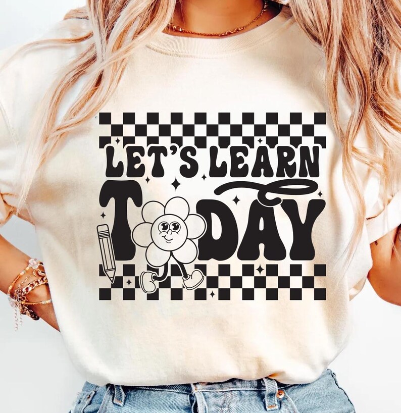 Op de afbeelding: Cr&egrave;mekleurig T-shirt met de tekst "LET'S LEARN TODAY" in een retro lettertype. Het ontwerp bevat een lachend bloemkarakter, een potlood en een dambordpatroon.