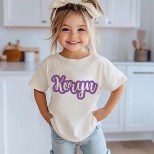 K&ouml;nnte beinhalten: Ein cremefarbenes T-Shirt mit dem Namen "Koryn" in lila Schrift. Das Kind tr&auml;gt eine blaue Jeans und eine cremefarbene Schleife im Haar. Der Hintergrund ist eine K&uuml;che mit wei&szlig;en Schr&auml;nken.