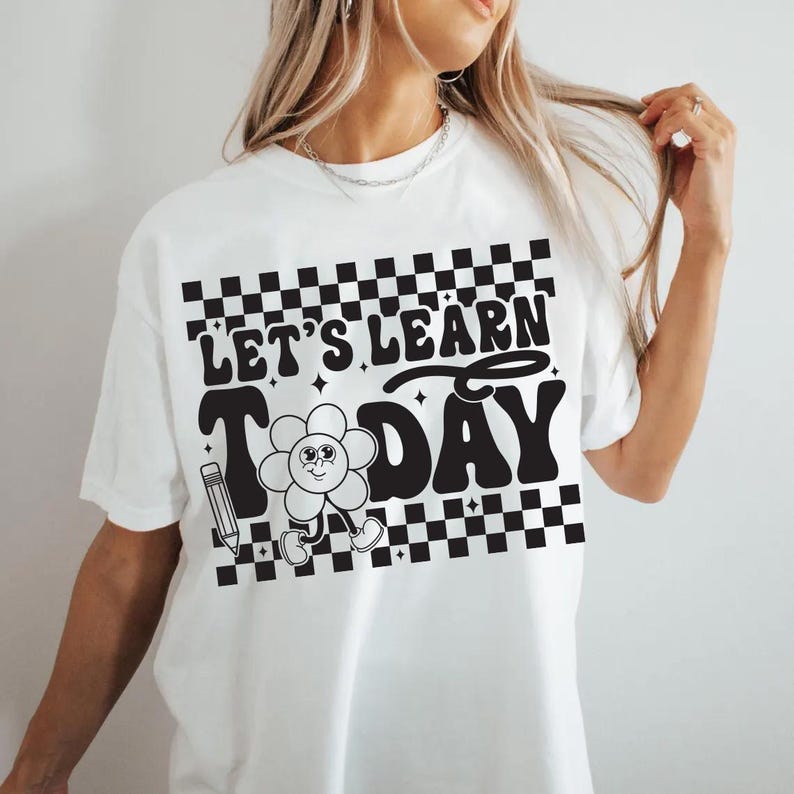Op de afbeelding: Wit T-shirt met een zwart-wit dambordontwerp aan de boven- en onderkant. Het shirt heeft de tekst "LET'S LEARN TODAY" in een retro lettertype, samen met een lachende bloem en een potloodafbeelding. Een leuk en educatief ontwerp.