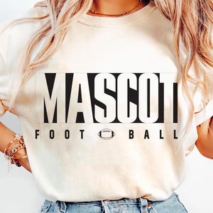 May include: Cream-colored sweatshirt with the word "MASCOT" in large, bold, black letters. Below the word "MASCOT" are the words "FOOT BALL" with a football graphic in between. The sweatshirt is a casual style.