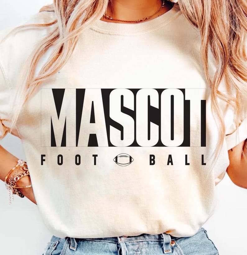 May include: Cream-colored sweatshirt with the word "MASCOT" in large, bold, black letters. Below the word "MASCOT" are the words "FOOT BALL" with a football graphic in between. The sweatshirt is a casual style.