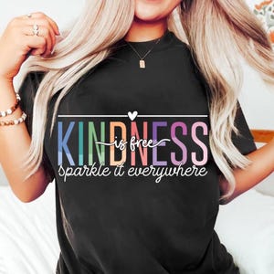 以下が含まれることがあります： 「KINDNESS is free sparkle it everywhere」というカラフルな文字がプリントされた黒いTシャツ。Tシャツはクルーネックで半袖です。テキストは青、緑、オレンジ、ピンク、紫など、さまざまな色で表示されています。