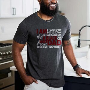 Op de afbeelding: Donkergrijs t-shirt met de woorden "I AM" in rood en "DAD" in rood, met andere woorden in wit, waaronder "LEADER", "BLACK" en "KING". Het shirt heeft korte mouwen en een ronde hals.