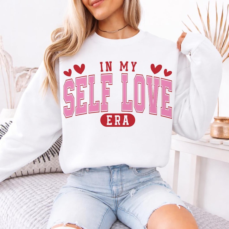 May include: White sweatshirt with the text "IN MY SELF LOVE ERA" in pink block letters with a red outline. The design includes red hearts above the text and a red oval with the word "ERA" at the bottom. The sweatshirt is a casual style.