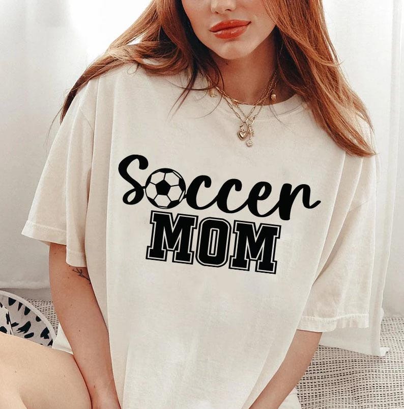 May include: White t-shirt with black text that says "Soccer Mom" and a black and white soccer ball graphic.