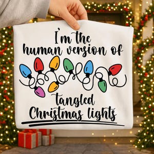 May include: A white t-shirt with the text "I'm the human version of tangled Christmas lights" in black. A string of colorful Christmas lights is above the text. The shirt is held up by a hand.
