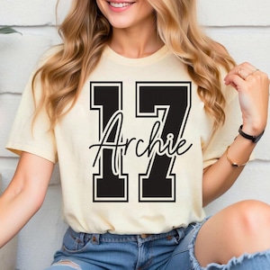 May include: A cream-colored t-shirt featuring the number "17" in a bold, black, collegiate-style font. The name "Archie" is scripted in black across the numbers. The shirt is short-sleeved.