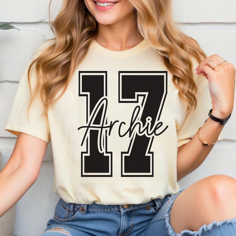 May include: A cream-colored t-shirt featuring the number "17" in a bold, black, collegiate-style font. The name "Archie" is scripted in black across the numbers. The shirt is short-sleeved.