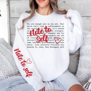 May include: White sweatshirt with the text "Note to Self" in red on the sleeve and chest. The chest features a heart graphic and a longer message in a typewriter font. A cozy and inspirational apparel item.