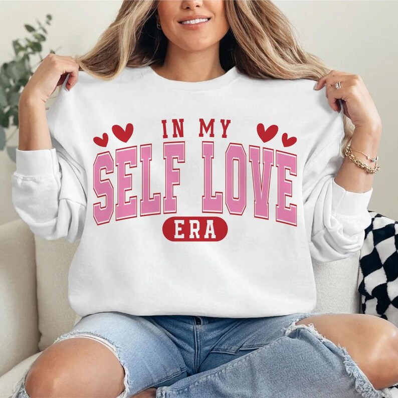May include: White sweatshirt with the text "IN MY SELF LOVE ERA" in pink block letters. The design includes red heart graphics above the text and a red oval with the word "ERA" at the bottom. The sweatshirt is a casual style.