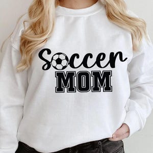May include: White sweatshirt with black text that reads "Soccer Mom" and a black and white soccer ball graphic.