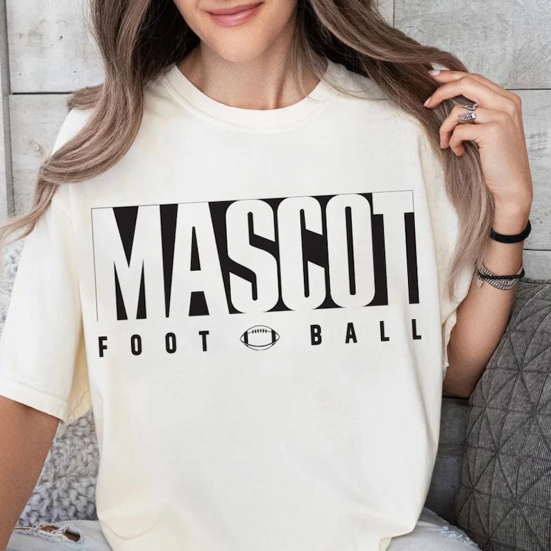 May include: Cream-colored t-shirt with the word "MASCOT" in bold black letters, above "FOOT BALL" and a football graphic. The shirt has a classic crew neck and short sleeves.