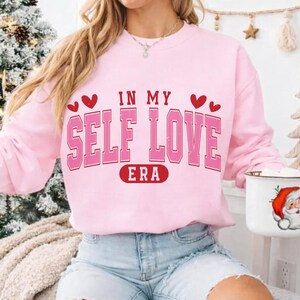 May include: A light pink sweatshirt with the text "IN MY SELF LOVE ERA" in a bold, pink, collegiate-style font. Red hearts accent the top of the text. The sweatshirt is a casual, comfortable style.
