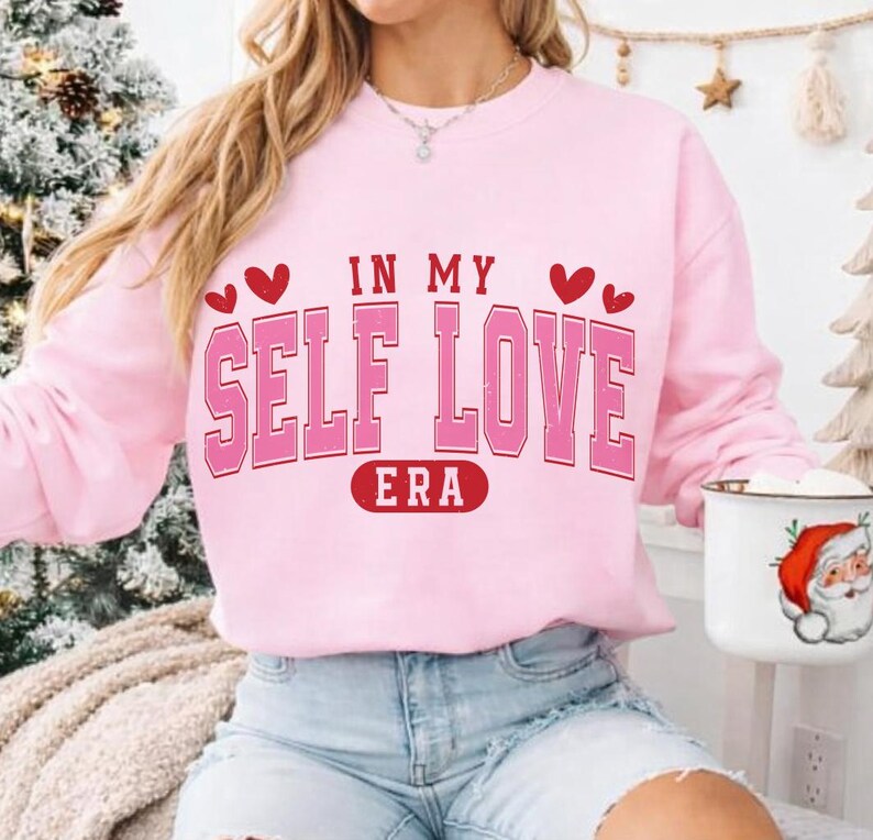 May include: A light pink sweatshirt with the text "IN MY SELF LOVE ERA" in a bold, pink, collegiate-style font. Red hearts accent the top of the text. The sweatshirt is a casual, comfortable style.