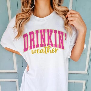 May include: White t-shirt with pink and gold lettering that reads "Drinkin' weather".