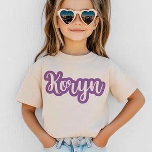 K&ouml;nnte beinhalten: Beiges T-Shirt mit dem Namen "Koryn" in lila Schrift. Das kurz&auml;rmelige Shirt wird von einer Person getragen, die herzf&ouml;rmige Sonnenbrillen tr&auml;gt.