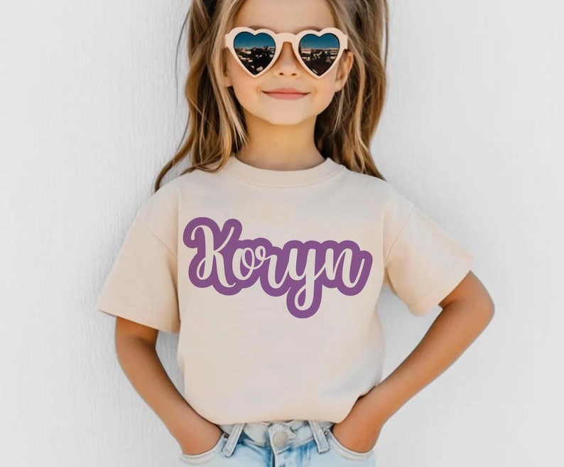 K&ouml;nnte beinhalten: Beiges T-Shirt mit dem Namen "Koryn" in lila Schrift. Das kurz&auml;rmelige Shirt wird von einer Person getragen, die herzf&ouml;rmige Sonnenbrillen tr&auml;gt.